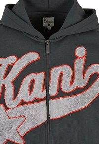 Sweat à capuche zippé gris foncé avec un grand logo texturé « Kani » souligné d’orange vif sur la poitrine et une étoile intégrée dans le texte.