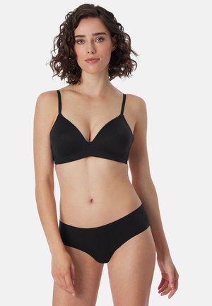 Kiharatukkainen nainen mustassa rintaliiveissä ja mustissa alushousuissa yksivärisen valkoisen taustan edessä.