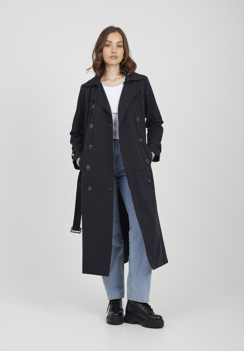 Czarny płaszcz trenchowy z podwójnym zapięciem, szerokim kołnierzem, paskiem i guzikami, w połączeniu z jasnoniebieskimi dżinsami i czarnymi botkami.