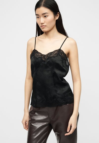 Ženska oblečena v črno majico z čipkastim robom in tankimi naramnicami ter temno rjave usnjene hlače, stoji pred enobarvnim belim ozadjem.