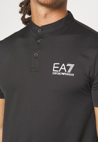 Melnā polo krekla ar strukturētu audumu, kuram ir īss stāvošs apkakle, trīs pogas un balts EA7 Emporio Armani logo uz krūtīm.