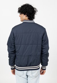 Marineblauwe bomberjack met een doorgestikt patroon, geribde kraag en manchetten met witte strepen, en een zakdetail op de mouw.