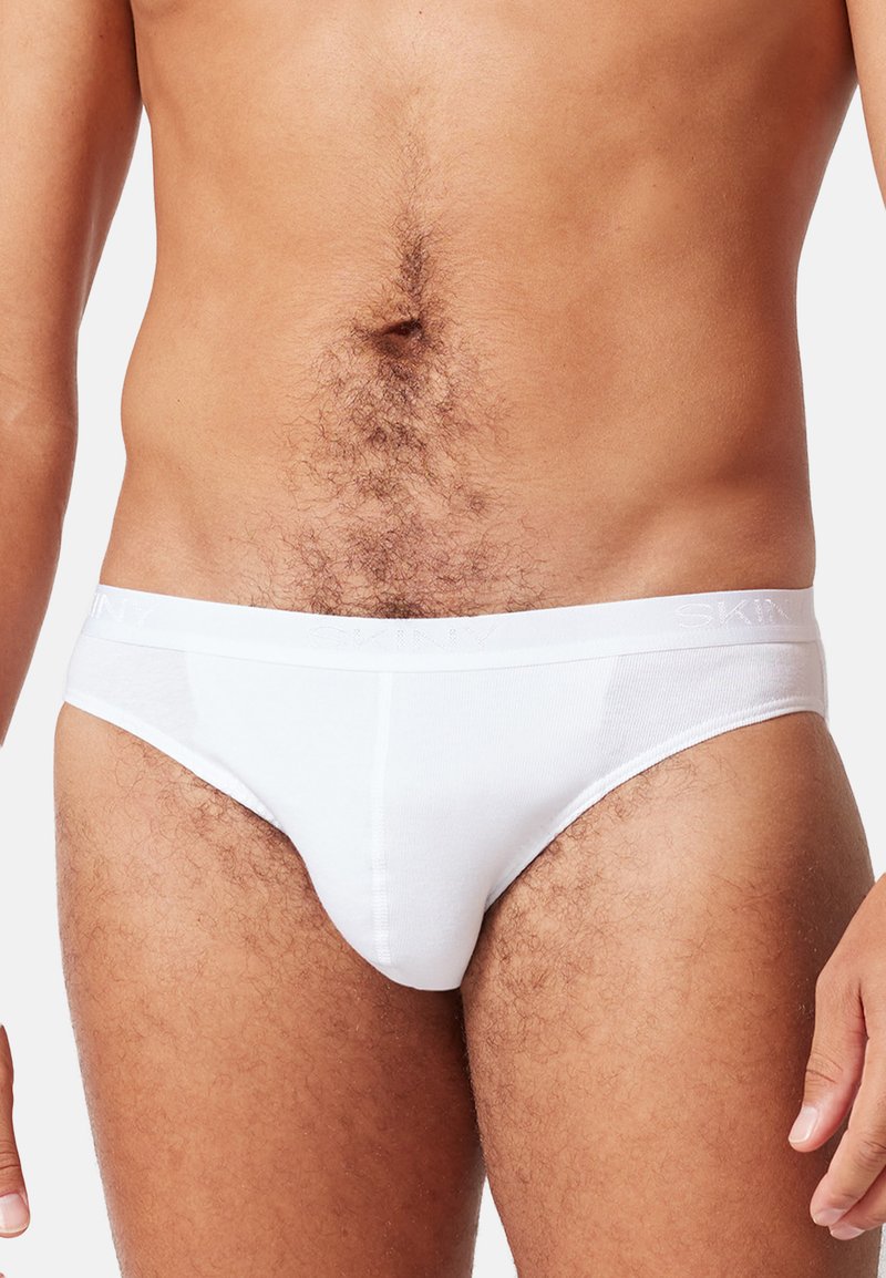 Mann in weißen Unterhosen mit einem gebrandeten Bund, der vor einem schlichten Hintergrund steht, wobei der Oberkörper und die Oberschenkel sichtbar sind.