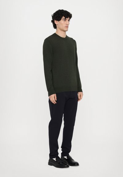 Dunkelgrüner, langärmeliger Pullover mit Rundhalsausschnitt, kombiniert mit maßgeschneiderten dunkelblauen Hosen und schwarzen Slippern. Glatte Stricktextur.