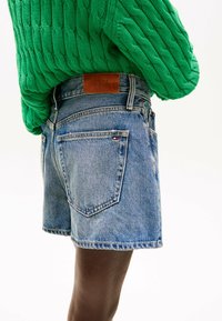 Shorts en denim à délavage moyen avec un ourlet brut, dotés de poches arrière et d'un patch en cuir. Portés avec un pull en maille texturée vert.