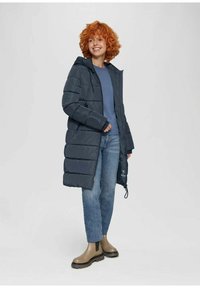 Dunkelgraue Pufferjacke mit Kapuze, vertikaler Steppung, Seitentaschen und verstellbarem Kordelzug am Saum. Getragen mit blauem Oberteil und Jeans.