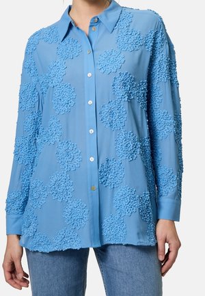 Femme portant une chemise bleu clair à manches longues avec des motifs floraux texturés, associée à un jean bleu.