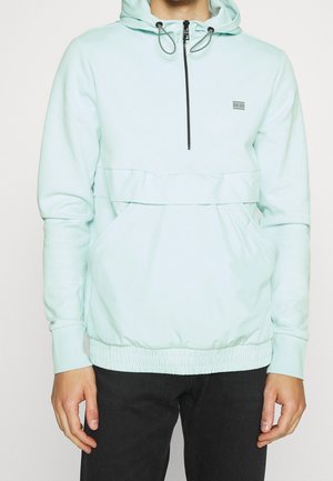 Hellblauer Kapuzenpullover mit vorderem Reißverschluss, Kängurutasche, elastischem Bund und schwarze Hose an einer Person vor weißem Hintergrund.