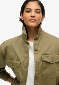 Khaki koszula z guzikami i wysokim kołnierzykiem, z dwiema kieszeniami na piersi oraz metką "SUPERDRY". Materiał wydaje się gładki; guziki są w kolorze dopasowanej zieleni.