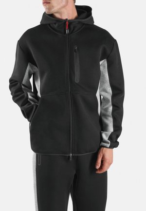 Homme portant un sweat à capuche noir zippé avec des panneaux gris sur les manches et les côtés, debout les mains partiellement dans les poches, tête tournée vers la gauche.