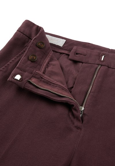 Burgundowe spodnie sztruksowe z zamkiem błyskawicznym, dwoma brązowymi guzikami i kieszonką na monety. Tkanina ma prążkowaną strukturę.
