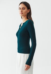 Teal long-sleeve top with a ribbed texture, featuring a slight V-neck and two buttons at the neckline. Fitted design, worn with white pants.