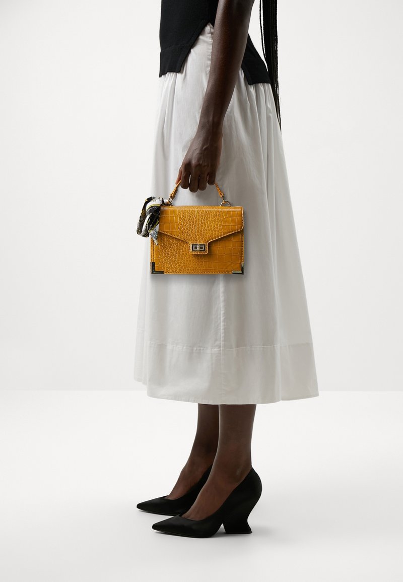 Personne portant un haut noir, une jupe midi blanche, des talons noirs, tenant un petit sac à main jaune texturé avec un foulard noué à la poignée.