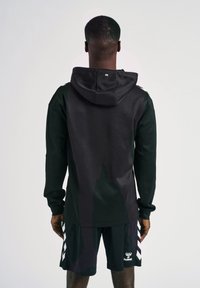 Sudadera negra con textura suave, que presenta una gran capucha y un logo en la parte posterior, combinada con pantalones cortos negros que exhiben un patrón de flechas blancas.