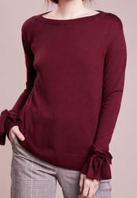 Maglione bordeaux realizzato in morbido tessuto a maglia, con scollatura ampia e lacci sulle maniche, abbinato a pantaloni a quadri.