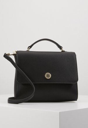 Sac à main en cuir noir texturé avec poignée supérieure et bandoulière amovible, présentant un petit fermoir rond doré avec logo sur le rabat avant.