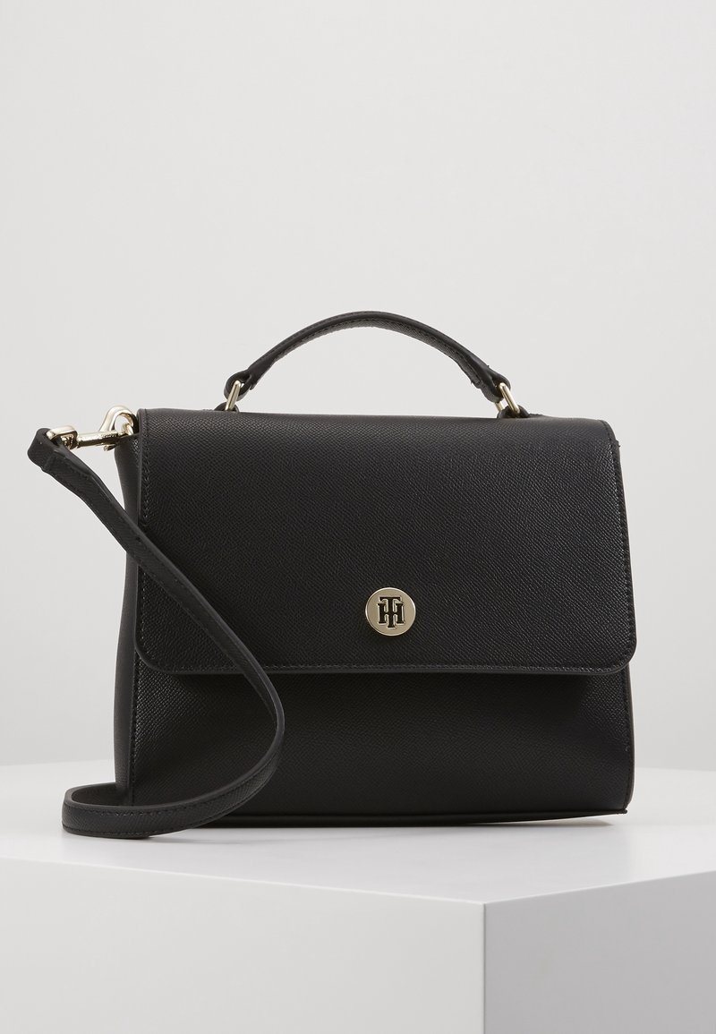 Sac à main en cuir noir texturé avec poignée supérieure et bandoulière amovible, présentant un petit fermoir rond doré avec logo sur le rabat avant.