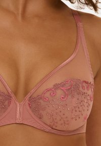 Lingerie rose clair de forme triangulaire. Présente une délicate broderie en rose plus foncé, des motifs floraux et des bretelles réglables.