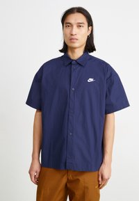 Tengeri kékszínű, rövid ujjú, gombos ing textúrázott kivitelben, fehér Nike logóval a mellrészen, barna nadrággal párosítva.