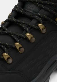 Zwarte wandelschoen met gestructureerd materiaal, goudkleurige hardwaredetails en zwarte veters met gele spikkels. Duurzaam ontwerp met een robuuste zool.