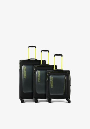 Drei schwarze American Tourister Koffer mit gelben Teleskopgriffen, Frontreißverschlusstaschen und vier Drehrollen, nach Größe angeordnet.