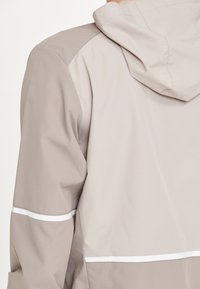 Ljust grå jacka med huva, som har kontrasterande paneler och vita detaljer. Slät textur med ett vattenavvisande ytfinish.