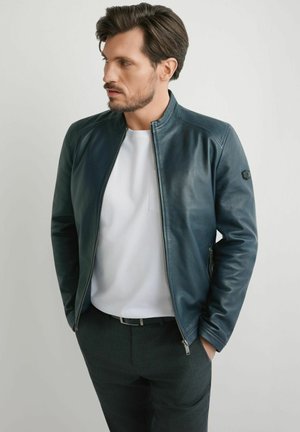 Mężczyzna w ciemnoniebieskiej skórzanej kurtce, białej koszuli i ciemnych spodniach stoi z rękami w kieszeniach, patrząc w lewo na jednolitym tle.