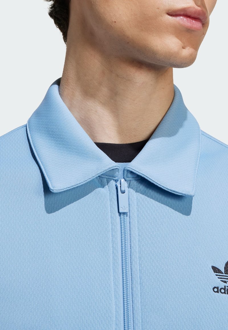 Veste zippée bleu clair en tissu texturé, avec un col rabattu et un petit logo noir sur le côté gauche près de l'encolure.