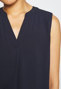 Marineblauwe mouwloze blouse met een V-hals, voorzien van subtiele stikseldetails en een gladde, lichte stof.