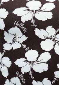 Μοτίβο λευκού λουλουδιού ιβίσκου με το κείμενο "Hollister" τυπωμένο επανειλημμένα σε σκουρό καφέ ύφασμα.
