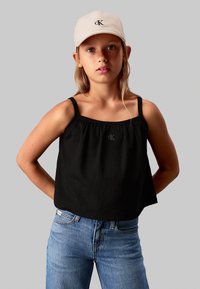 Top negro sin mangas con textura fruncida, que presenta un pequeño logo bordado "cK" en la parte frontal, combinado con jeans de mezclilla azules.