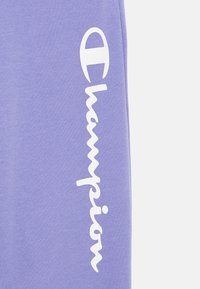 Μωβ φόρμα από απαλό, υφασμάτινο ύφασμα με λευκό λογότυπο και όνομα μάρκας τυπωμένα κάθετα κατά μήκος της πλευράς.