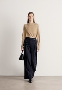 Beżowa bluzka z długim rękawem oraz granatowe, wysokie spodnie z plisami, ozdobione złotymi guzikami i małą czarną torebką. Gładkie faktury.