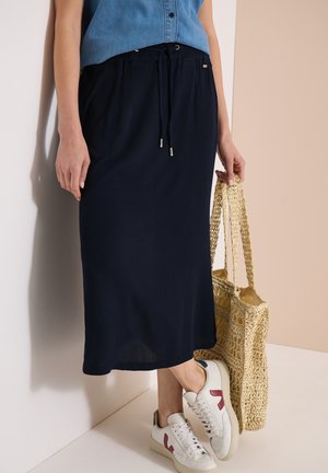 Femme portant une jupe midi bleu marine avec cordon, des baskets blanches avec un logo bordeaux, et tenant un sac fourre-tout beige tissé.