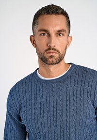 Blauer gestrickter Pullover mit einem Kabelmuster, Rundhalsausschnitt und strukturiertem Stoff. Über einem weißen Hemd getragen, mit einer Nahaufnahme.