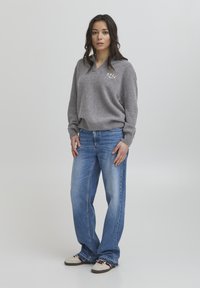 Suéter gris de punto con escote en V y el texto 'BALL'. Combinado con jeans de pierna ancha de color azul claro y zapatillas beige con acentos oscuros.
