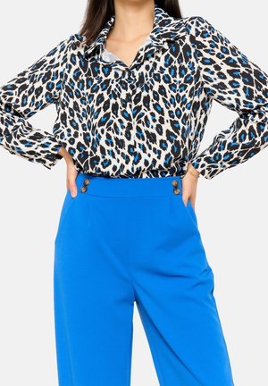 Frau, die ein langärmliges weißes Hemd mit blauem und schwarzem Leopardenmuster trägt, das in eine hoch taillierte, leuchtend blaue Hose mit Knöpfen gesteckt ist.