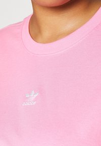 Närbild av en person som bär en ljusrosa Adidas t-shirt med en vit Adidas-logotyp centrerad under halsringningen.
