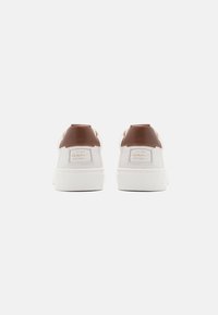 White sneakers with a brown leather heel section, featuring a textured sole and branding on the back in gold lettering. Clean and minimalist design.