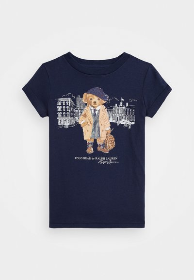 T-shirt de algodão azul-marinho apresentando um urso em um casaco e boné bege, segurando uma bolsa, com uma ilustração detalhada de uma paisagem urbana abaixo.