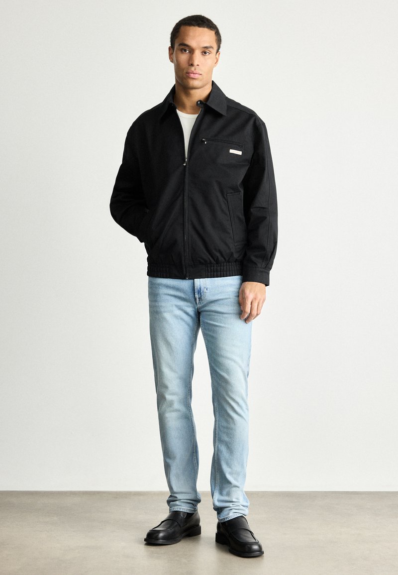 Black zip-up jacket with a collar, front pocket, and elastic hem; paired with light blue jeans and black shoes on a neutral background.