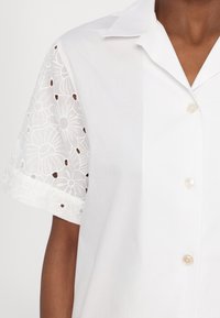 Chemise blanche à manches courtes avec motif en dentelle florale sur les manches et boutons nacrés portée par une personne à la peau de ton moyen-foncé.