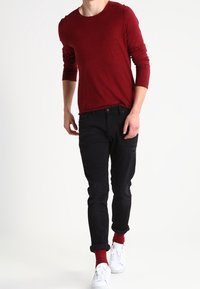Rotes Langarmhemd, schwarze schmal geschnittene Jeans, weiße Sneakers und bordeauxfarbene Socken. Einfaches Design mit lockerer Passform und hochgerollten Säumen an den Jeans.