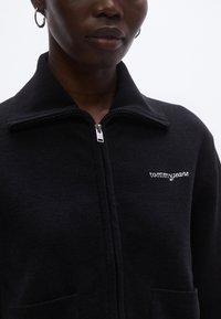 Fekete, cipzáras dzseki puha anyagból, magas gallérral, elülső zsebekkel és bal oldali mellrészen hímzett "tommyjeans" logóval.