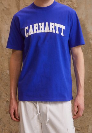 Person mit leuchtend blauem Carhartt-T-Shirt und weißen Hosen mit Kordelzug vor einem strukturierten beigen Hintergrund.