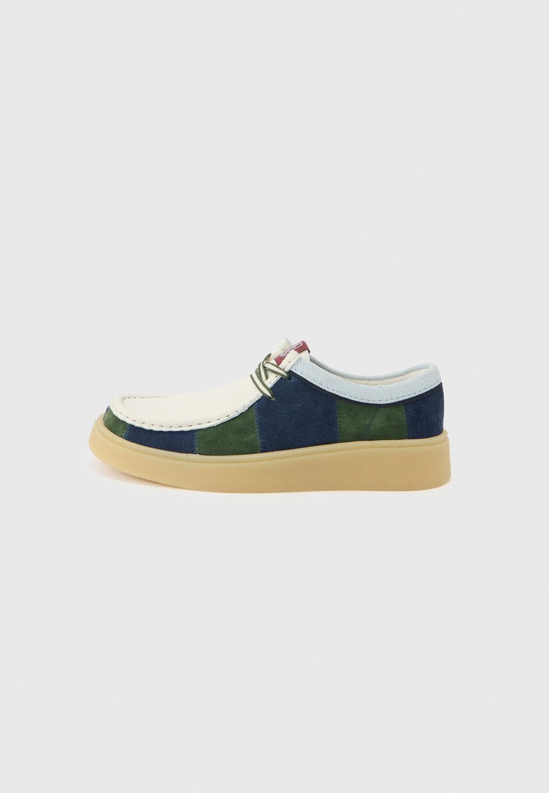 Chaussure décontractée basse avec bout crème, panneaux en daim bleu et vert, lacets blancs et une semelle épaisse en caoutchouc beige sur un fond uni.