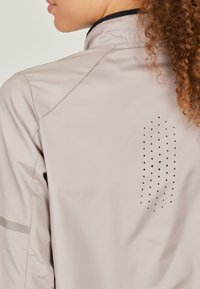 Light beige jacket featuring a high collar, smooth fabric, and a perforated design on the back. Reflective accents are also present.