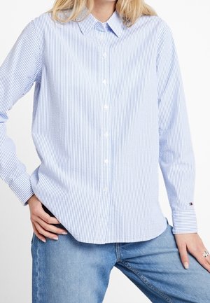 Chemise à rayures bleues et blanches avec un col et des manches longues, présentant une coupe droite et des accents de marque subtils au niveau des poignets.