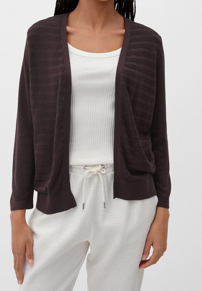Femme portant un cardigan marron foncé ouvert devant, sur un top blanc côtelé et un pantalon blanc texturé à cordon de serrage.