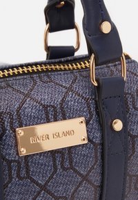 Borsa strutturata di colore blu scuro e in tessuto texturizzato con un'etichetta in metallo dorato. Presenta una chiusura con zip e due manici per il trasporto.